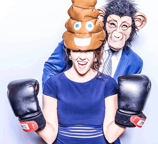 Two people in a photo booth, one wearing a poop emoji hat and boxing gloves, the other wearing a monkey mask