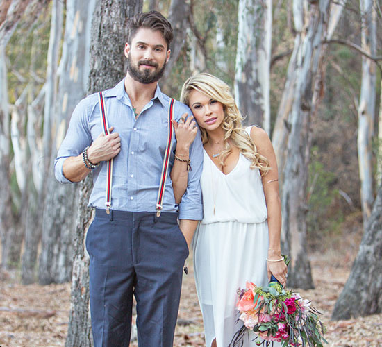 Couple posing outdoors, the man wearing suspenders and a shirt, the woman in a white dress holding a bouquet of flowers