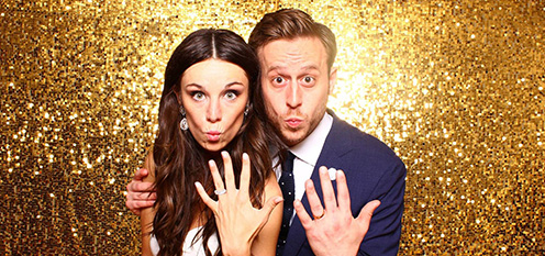 Couple posing with their hands showing rings, in front of a gold glittery backdrop