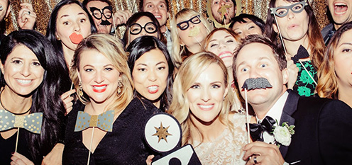 Large group of people posing with photo booth props, including glasses, mustaches, and bow ties, in front of a gold backdrop